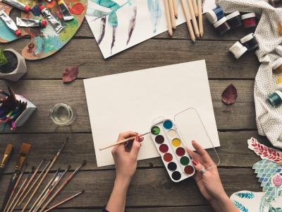 Viser et bord med materialer til tegning og maling og en hånde der maler med vandfarver